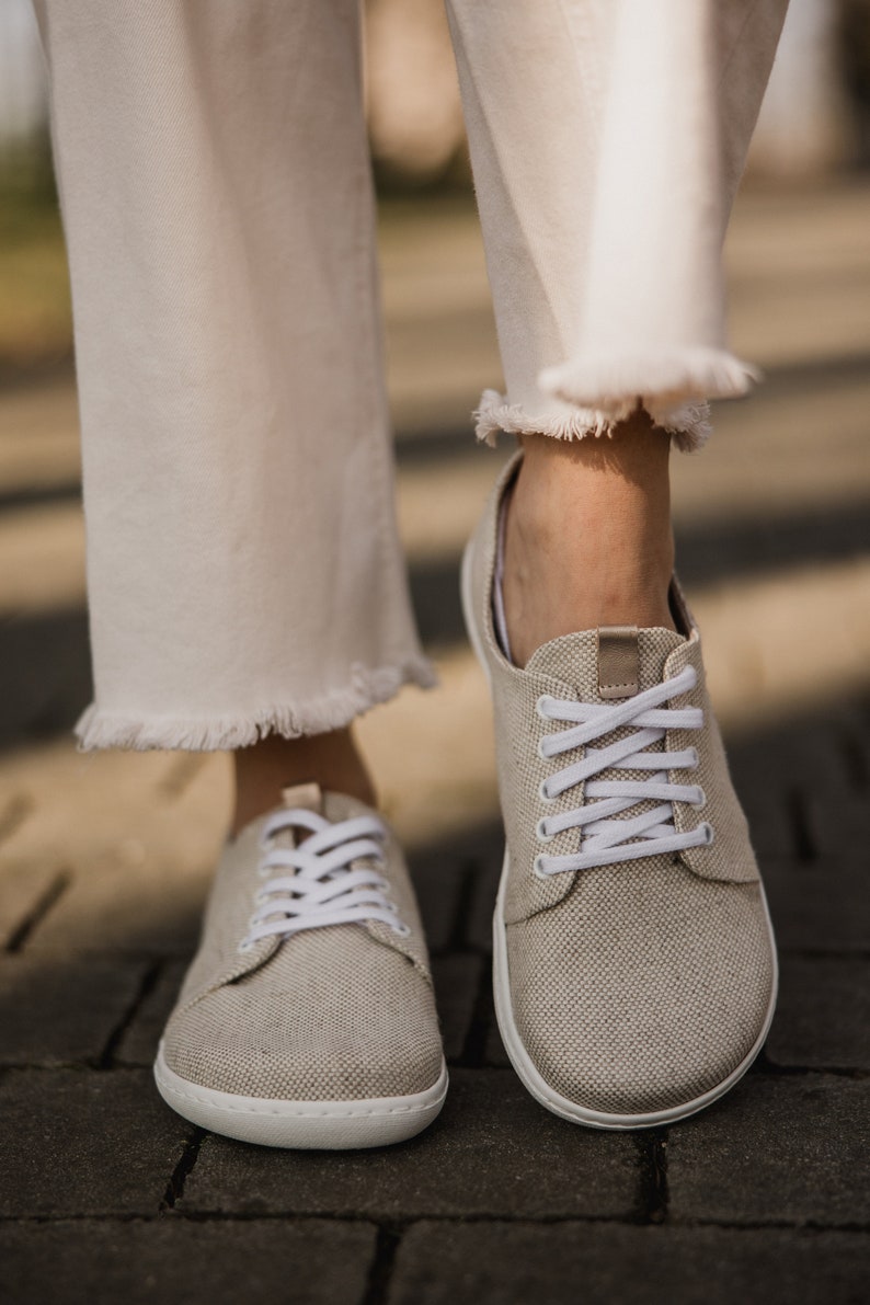 May include: A pair of beige canvas sneakers with white laces. The sneakers have a white rubber sole and are worn by a person wearing white pants.