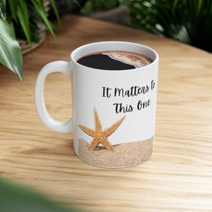 May include: White ceramic coffee mug with a black handle. The mug has a starfish and sand design and the text "It Matters to This One".