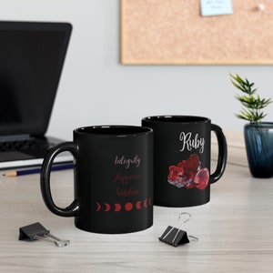 May include: Two black ceramic mugs. One mug features the word "Ruby" in white script with a red gemstone illustration. The other mug has the words "Integrity, Happiness, Wisdom" in a vertical layout, with red moon phase graphics.