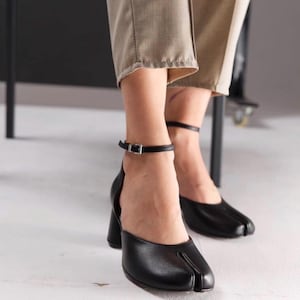 May include: Black leather heeled shoes with a split-toe design. The shoes have a thick heel and an ankle strap with a silver buckle. The shoes are worn with beige trousers.