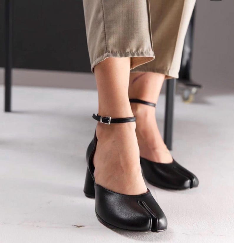 May include: Black leather heeled shoes with a split-toe design. The shoes have a thick heel and an ankle strap with a silver buckle. The shoes are worn with beige trousers.