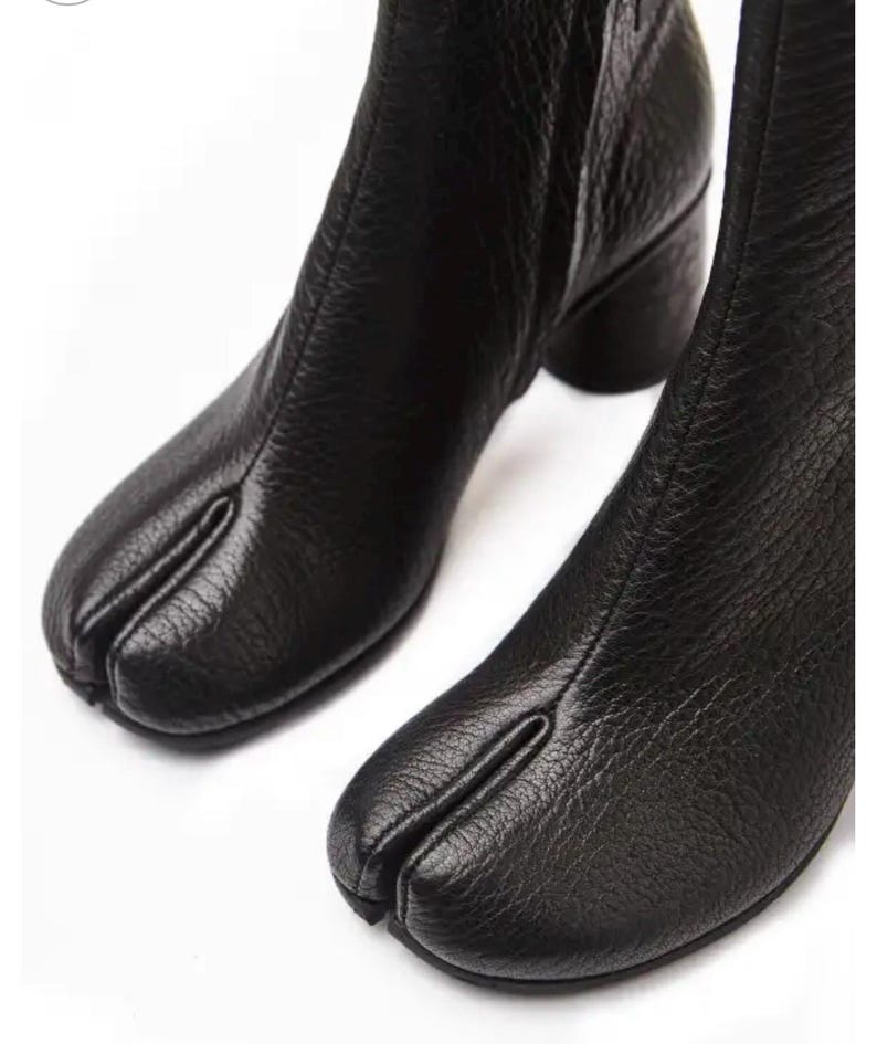 May include: Two black leather ankle boots with a split-toe design. The boots have a textured finish and a low, block heel. The split-toe design is a distinctive feature. The boots are set against a white background.