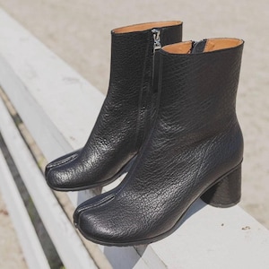 May include: A pair of black leather ankle boots with a unique split-toe design. The boots feature a textured surface, a side zipper, and a block heel. The boots are displayed on a white surface.
