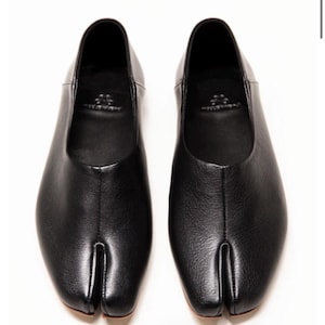 May include: A pair of black leather split-toe flats. The shoes feature a rounded toe and a split design separating the big toe. The shoes are presented on a white backdrop.