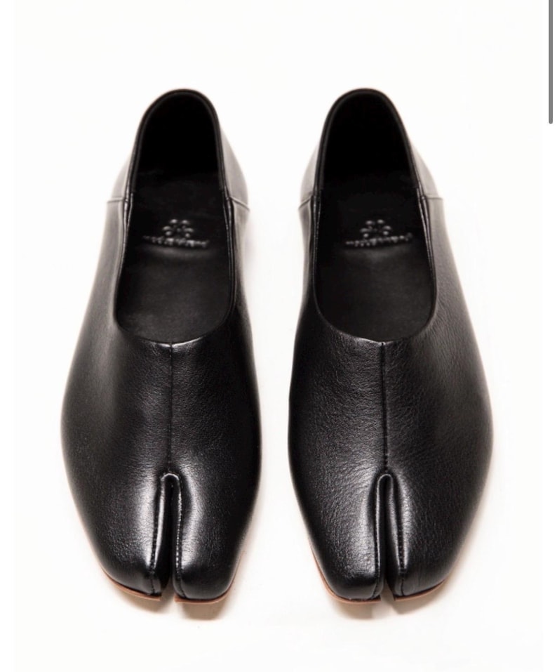 May include: A pair of black leather split-toe flats. The shoes feature a rounded toe and a split design separating the big toe. The shoes are presented on a white backdrop.