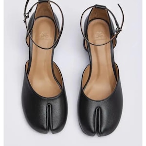 May include: A pair of black leather Tabi shoes with ankle straps. The shoes feature a split-toe design and a small heel. The ankle straps are secured with a small buckle. The shoes are displayed on a white background.