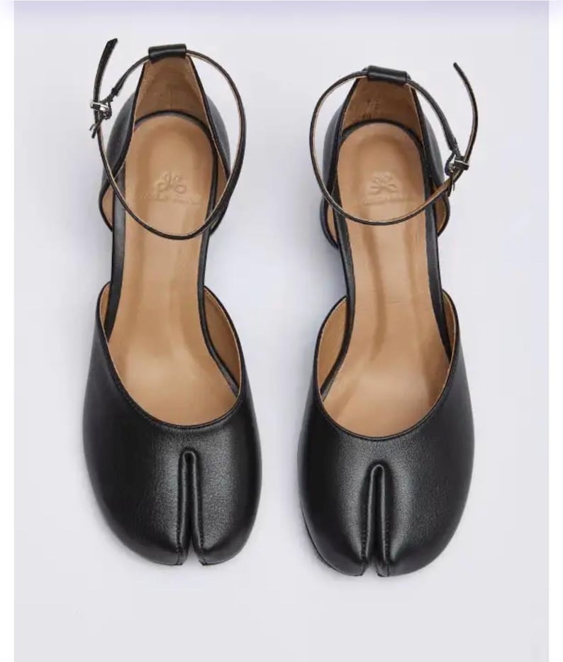 May include: A pair of black leather Tabi shoes with ankle straps. The shoes feature a split-toe design and a small heel. The ankle straps are secured with a small buckle. The shoes are displayed on a white background.