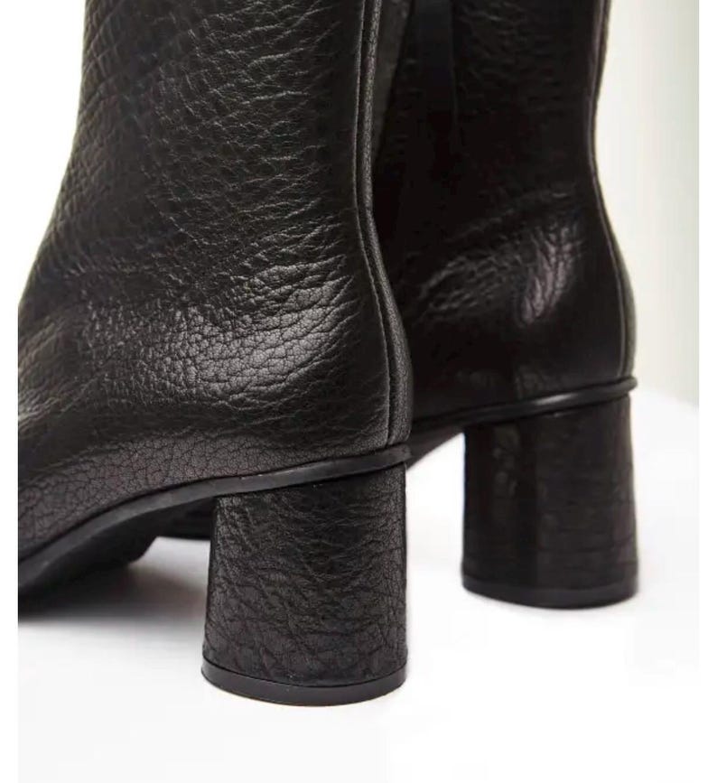 May include: Close-up of two black leather ankle boots. The boots have a textured surface and a chunky, block heel. The heels appear to be approximately 7.6 cm tall. The boots are set against a white background.