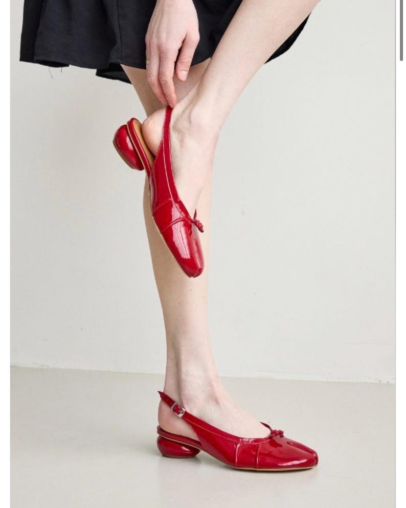 May include: Red patent leather slingback flats with a small bow detail on the toe. The shoes have a rounded heel and a buckle closure. The style is a modern take on a classic design.