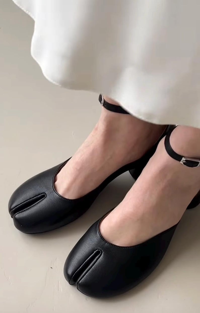 May include: Black leather split-toe shoes with a small heel and ankle strap. The shoes have a unique design with a separate section for the big toe. The ankle straps have a small silver buckle.