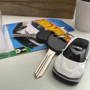 Puede incluir: Un coche de juguete blanco con una llave negra y una tarjeta de coche Hot Wheels. La tarjeta de coche tiene un fondo azul con el logotipo de Hot Wheels y un coche eléctrico blanco.