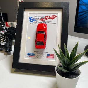 May include: A framed display of a red 1983 Ford Mustang 3rd generation, 5.0 liter hatchback. The display includes the Ford Performance logo, the American flag, and the car's specifications: Horsepower 225 hp, Torque 300 lb-ft, Compression 9.0:1, 0 to 60 mph 7.0 seconds, Top speed 124 mph.