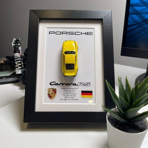 May include: A framed display featuring a yellow toy car model of a Porsche 911 Carrera RS 2.7. The frame includes the car's name, specifications, and a German flag.
