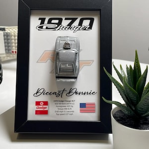 Peut inclure: Une vitrine encadrée d'une miniature de voiture Dodge Charger R/T argentée de 1970. Le cadre a un fond noir avec du texte blanc qui dit "1970 Charger" et "Diecast Donnie". Le cadre comprend également le logo Dodge et le drapeau américain. Le texte sous la voiture énumère les spécifications de la voiture, y compris la taille du moteur, la puissance, le couple, le temps de 0 à 100 km/h et la vitesse maximale.