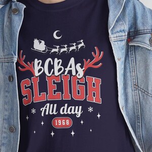 May include: Navy blue t-shirt with a festive design. The shirt features the text "BCBAs SLEIGH All day 1968" in red and white, along with a Santa sleigh and reindeer graphic. A crescent moon and stars complete the holiday theme.