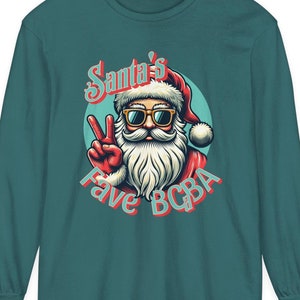 Puede incluir: Una camiseta de manga larga de color turquesa con una imagen de dibujos animados de Papá Noel con gafas de sol y haciendo un signo de paz. El texto "Santa's Fave BCBA" está impreso debajo de la imagen.