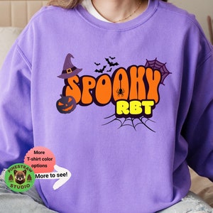 Puede incluir: Sudadera morada con un diseño gráfico de un sombrero de bruja, murciélagos, una telaraña, una calabaza y el texto "Spooky RBT".