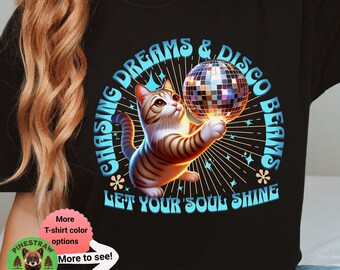 May include: A black t-shirt with a graphic of a cat chasing a disco ball. The text "Chasing Dreams & Disco Beams" is written in a retro font around the cat and disco ball. The text "Let Your Soul Shine" is written below the cat and disco ball.