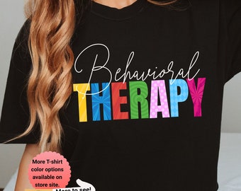 May include: Black t-shirt with the text "Behavioral THERAPY" in rainbow colours. The text is stylised with different patterns in each letter. A small pink circle with white text says "More T-shirt colour options available on store site." A yellow arrow points to the right. A small brown circle with a bear illustration says "Pinestraw Studio Elevating the Everyday."