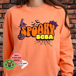 Puede incluir: Camiseta de manga larga naranja con un diseño gráfico de un sombrero de bruja, una telaraña, murciélagos, una calabaza y el texto "Spooky BCBA".