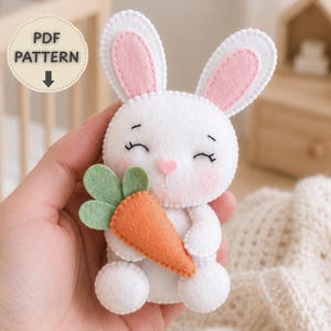 Pode incluir: Um peluche de coelho em feltro branco com interior das orelhas rosa, traços faciais bordados e bochechas rosa. O coelho segura uma cenoura de feltro laranja com folhas verdes. Um emblema circular exibe o texto PDF PATTERN.