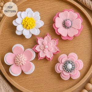 Pode incluir: Cinco flores de feltro feitas à mão em tons de branco, amarelo e vários rosas. As flores apresentam pétalas em camadas, centros texturizados e um pequeno detalhe de pérola branca. Um selo circular no canto superior esquerdo diz PDF PATTERN.