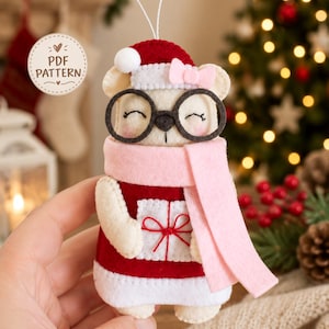 Può includere: Un ornamento in feltro a forma di orso che indossa un cappello da Babbo Natale rosso con un pon-pon bianco, una sciarpa rosa chiaro e occhiali tondi neri. L'orso ha un piccolo fiocco rosa sull'orecchio e tiene un regalo bianco. Testo: PDF PATTERN.