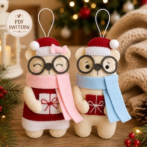 Pode incluir: Dois enfeites de feltro em forma de urso com laços para pendurar. Um urso usa um gorro vermelho, cachecol rosa e segura um presente branco. O outro usa gorro vermelho, cachecol azul e segura um presente vermelho. Um emblema diz PDF PATTERN.