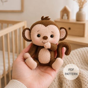 May include: A small, handcrafted felt monkey toy featuring dark brown and light beige fabric sections, black bead eyes, and a curled tail. The monkey has a stitched face with pink blush accents and is labeled with the text PDF PATTERN.