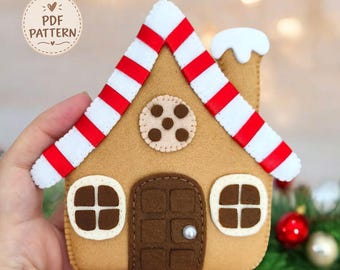 Puede incluir: Un adorno de fieltro con forma de casa de jengibre que mide aproximadamente 12 cm de altura. La casa presenta un techo a rayas rojas y blancas, una puerta marrón con un pomo perlado, dos ventanas, un detalle de galleta circular y una chimenea blanca.