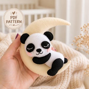 May include: A felt craft project featuring a sleeping panda bear resting on a cream-colored crescent moon. The panda has black ears, eye patches, and limbs with white fur and pink cheeks. A circular badge in the corner displays the text PDF PATTERN.