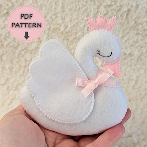 Può includere: Un cigno in feltro bianco con una corona e un fiocco rosa. Il cigno ha un collo curvo e un piccolo becco rosa. La corona è adornata da piccole perline d'argento. L'immagine include anche un cerchio rosa con il testo "PDF PATTERN" e una freccia verso il basso.