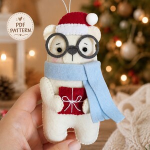 Pode incluir: Um ornamento de feltro de urso polar usando um gorro de Papai Noel vermelho com um pompom branco, óculos redondos pretos e um cachecol azul claro. O urso segura um pequeno presente vermelho com detalhes de fita branca. Rótulo PDF PATTERN.