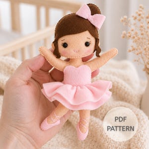 Peut inclure: Une poupée ballerine en feutrine avec des cheveux bruns ornés d'un nœud rose, un justaucorps rose clair, un tutu rose à volants et des chaussons de danse avec des lacets croisés. Yeux en perles noires et détails brodés. Texte PDF PATTERN inclus.