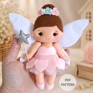 Pode incluir: Uma boneca fada de feltro com cabelo castanho, vestido rosa e asas brancas. A boneca apresenta uma coroa de flores com pequenas pérolas, sapatilhas de ballet rosa e segura uma varinha de estrela com glitter prateado. Texto PDF PATTERN.