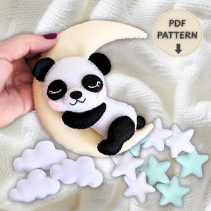 Pode incluir: Um projeto de artesanato em feltro com um urso panda a dormir sobre uma lua crescente de cor creme. O conjunto inclui duas nuvens de feltro branco e sete pequenas estrelas em branco e verde menta pálido. Uma etiqueta mostra PDF PATTERN.