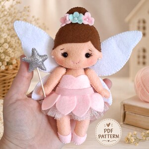 Puede incluir: Una muñeca hada de fieltro con cabello castaño, vestido rosa con falda de pétalos y alas blancas. La muñeca sostiene una varita de estrella con purpurina plateada y lleva una corona de flores. Una etiqueta circular muestra PDF PATTERN.