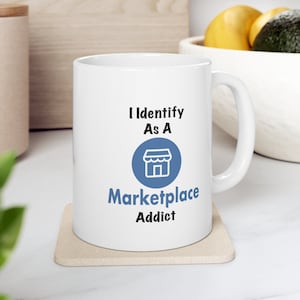 Könnte beinhalten: Weiße Keramik-Tasse mit dem Text "I Identify As A Marketplace Addict". Der Text ist in Schwarz und Blau gehalten, mit einem blauen Kreis und einem Shop-Symbol. Die Tasse steht auf einem beigen Untersetzer.