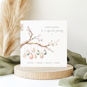 May include: Happy Easter to a special family card featuring a watercolour illustration of a tree branch with pink blossoms and hanging Easter eggs. The eggs are decorated with pastel colours and patterns. The names Stacey, Adam, Lillie, and Lydia are written below the branch.