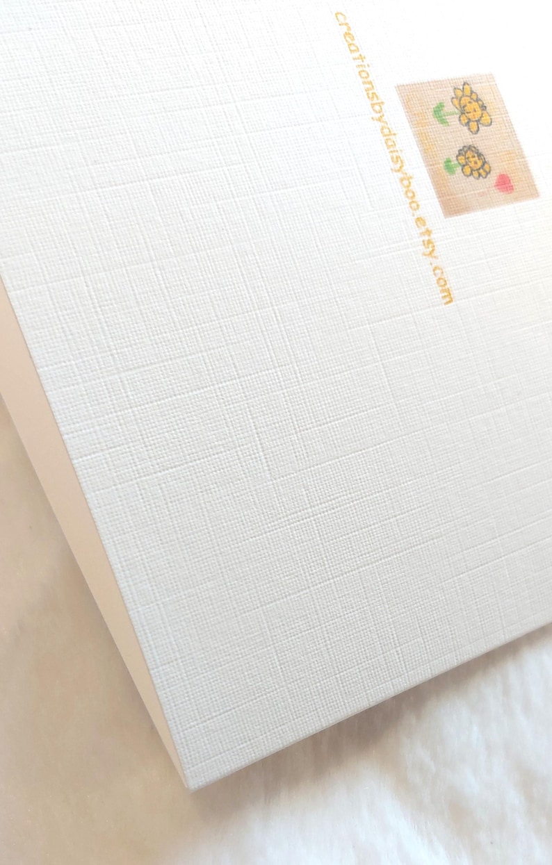 May include: White card with a textured surface. The back of the card has a small image of three yellow flowers and a red heart. The text "creationsbydaisyboo.etsy.com" is printed in gold on the back of the card.