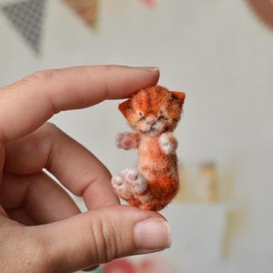Puede incluir: Una figura miniatura de gatito atigrado naranja hecho con fieltro de aguja, con rayas de color naranja oscuro, orejas pequeñas y puntiagudas y patas blancas diminutas. El gatito está sentado con las patas delanteras levantadas y textura suave.