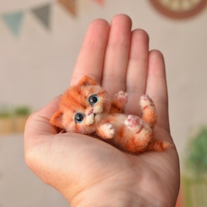 May include: A miniature needle-felted orange tabby kitten figurine with large blue eyes, pointed ears, and small paws, lying on its back. The kitten features a textured, fuzzy surface with darker orange stripes and a light-coloured muzzle.