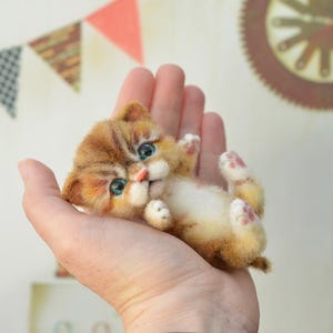 Op de afbeelding: Handgemaakte, oranje en witte kattenfiguur met blauwe ogen, liggend op zijn rug in een hand. Het katje heeft een zachte textuur en roze voetzolen. Decoratieve vlaggetjes en een klok zijn op de achtergrond.