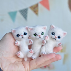 Peut inclure: Trois figurines de chatons blancs faits à la main, avec des yeux bleus et verts et des coussinets roses. Les chatons sont faits d'une matière douce et sont posés dans une posture ludique. L'arrière-plan comprend des guirlandes colorées.