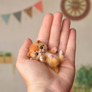 Può includere: Una statuina di gattino arancione in miniatura con occhi verdi e cuscinetti rosa è tenuta in una mano aperta. Il gattino è sdraiato sulla schiena. Lo sfondo include un muro con uno striscione decorativo e un orologio di legno.
