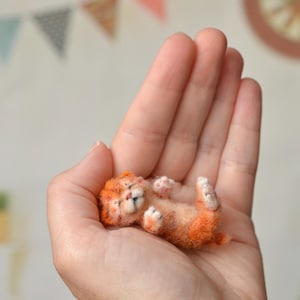 Peut inclure: Une figurine miniature de chaton tigré orange en laine feutrée, allongée sur le dos les yeux fermés. Cette petite sculpture de chat duveteux présente une fourrure texturée orange et crème, de minuscules coussinets roses et une queue recourbée.