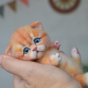 Puede incluir: Una pequeña figura de gatito atigrado naranja hecha con fieltro de aguja, con ojos azul brillante, nariz rosa y patas blancas. El gato en miniatura tiene una textura suave y esponjosa y se muestra en posición reclinada con las patas visibles.