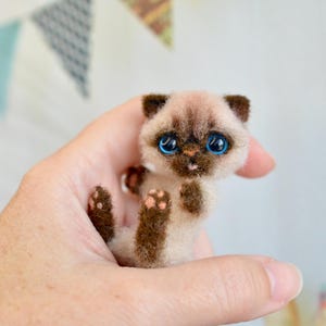 Puede incluir: Una figura en miniatura de gato siamés con cuerpo marrón claro, orejas y patas marrones oscuras y ojos azul brillante. El gatito artesanal se sostiene en una mano, mostrando su pequeño tamaño y detalles. El fondo incluye banderas triangulares de colores.