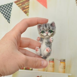 Może przedstawiać: Miniaturowa figurka szarego i białego kociaka z zielonymi oczami i różowymi poduszkami łap. Mały kotek trzymany jest w dłoni, prezentując swoje szczegółowe cechy i realistyczny wygląd. Figurka jest uroczym przykładem ręcznie robionej sztuki.