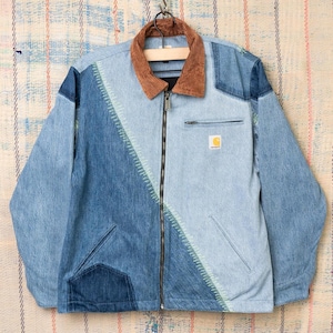 Peut inclure: Veste en jean patchwork avec un col en velours côtelé marron. La veste présente une fermeture éclair, plusieurs nuances de denim bleu et un petit logo carré jaune sur la poche poitrine. La veste est suspendue à un crochet en métal.